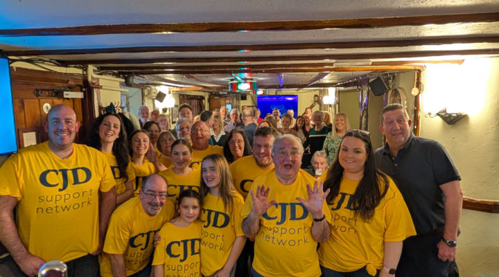 A group of people are standing together in a historical pub with ceiling timbers showing. They are wearing bright yellow 'CJD Support Network' t shirts and look happy. All ages are represented.