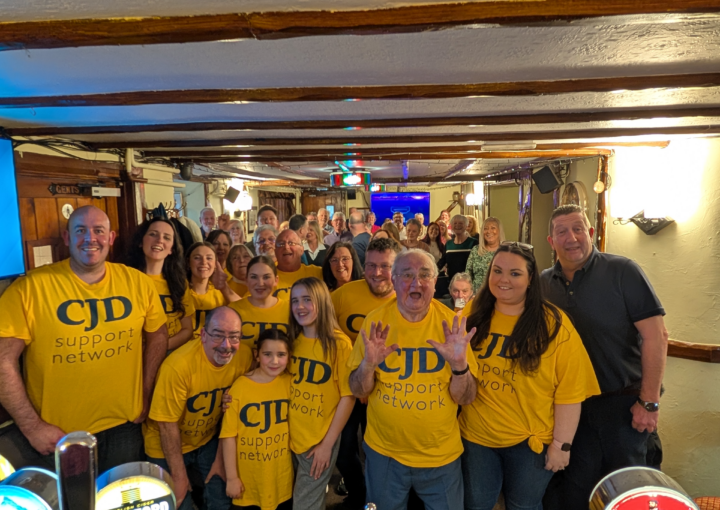 A group of people are standing together in a historical pub with ceiling timbers showing. They are wearing bright yellow 'CJD Support Network' t shirts and look happy. All ages are represented.