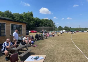 Onlookers are sitting on the grass at the side of a cricket pitch watching the game on a beautiful summer's day.