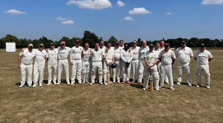 A cricket team stands proudly in their white uniforms on a cricket pitch. The sky is bright blue.