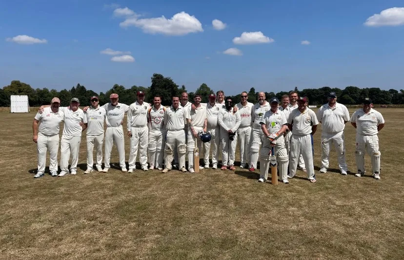 A cricket team stands proudly in their white uniforms on a cricket pitch. The sky is bright blue.